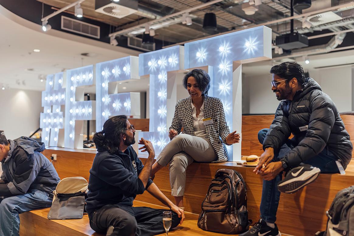 A group of smiling event attendees are sitting and chatting in front of a large #FEIT sign.