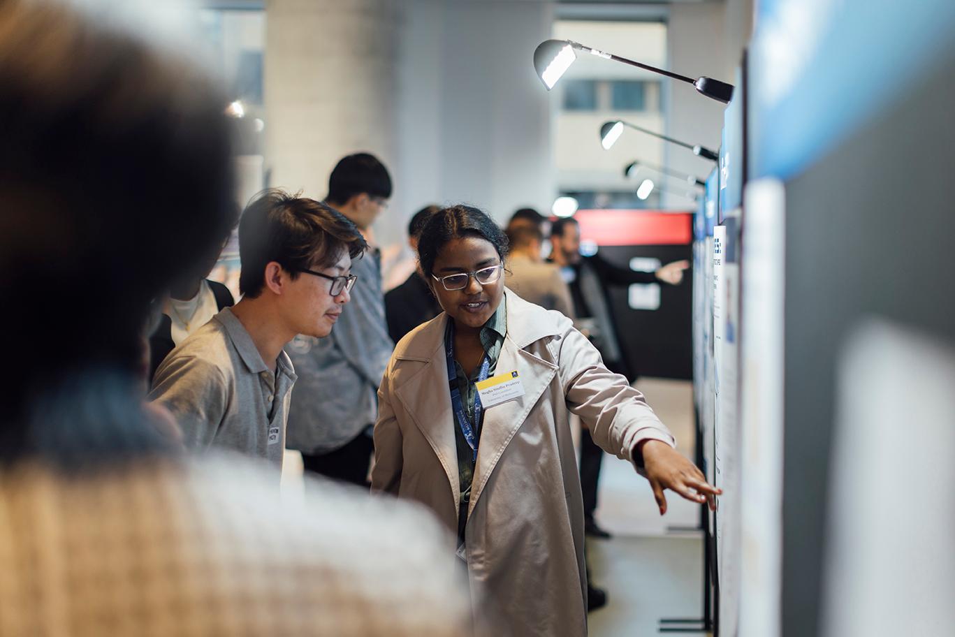 A graduate researcher explains her projects to an attendee while pointing to a poster board.
