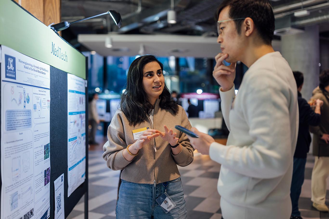 A graduate researcher explains her MedTech project to an attendee in front of a poster board.