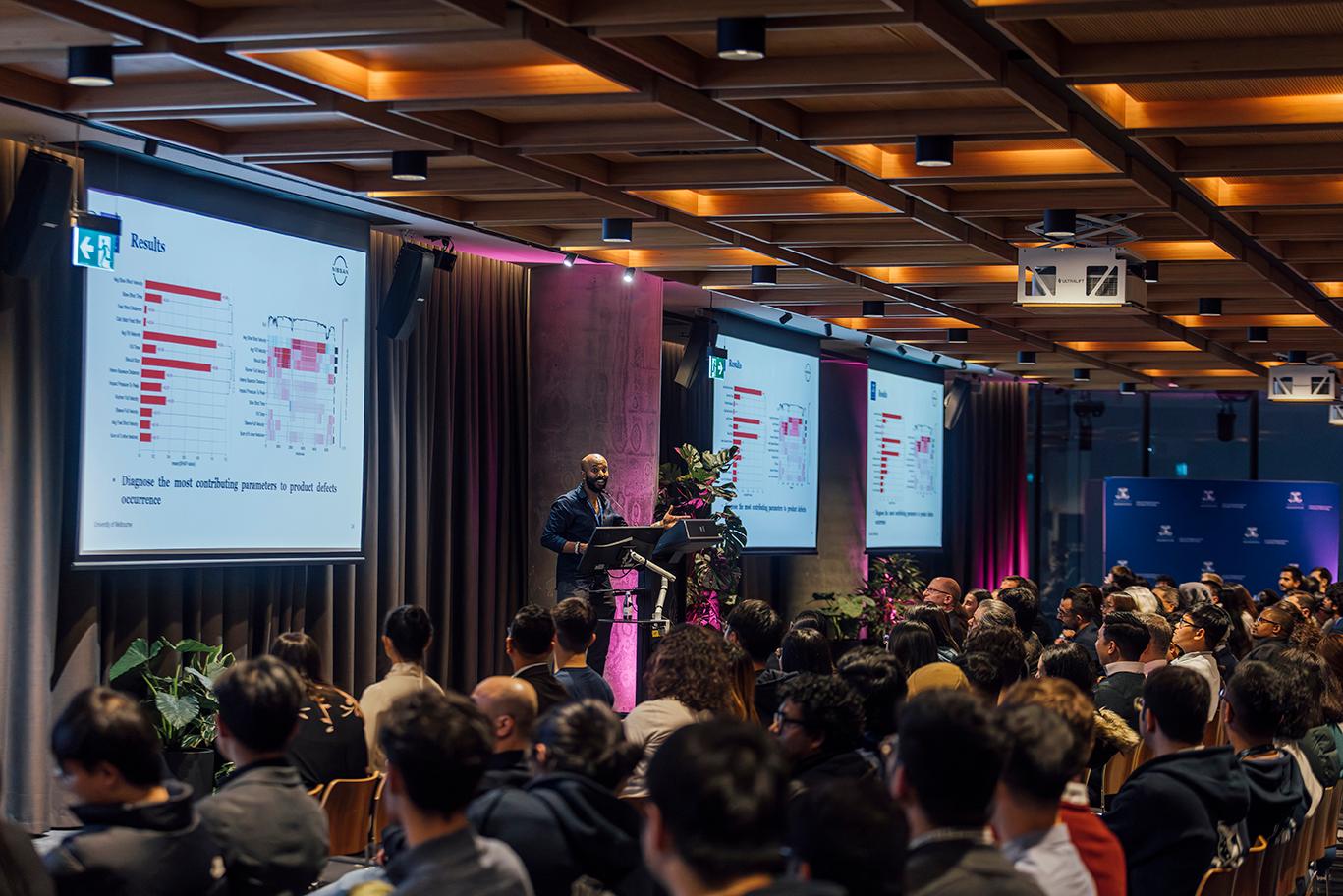 A male presenter is speaking to an audience, with three screens displaying graphs behind him.